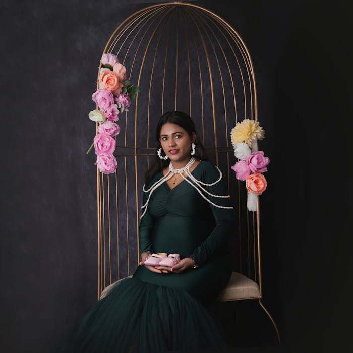 Pregnant woman in elegant dress holding baby shoes, seated in floral-decorated cage.