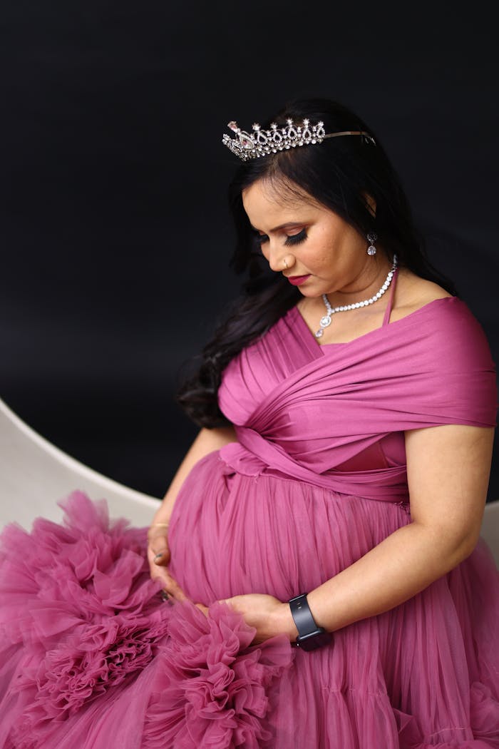A pregnant woman in a pink gown and tiara poses gracefully for a maternity portrait.