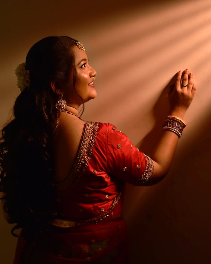 Back view of a smiling Indian woman in a red saree, adorned with jewelry, standing indoors.