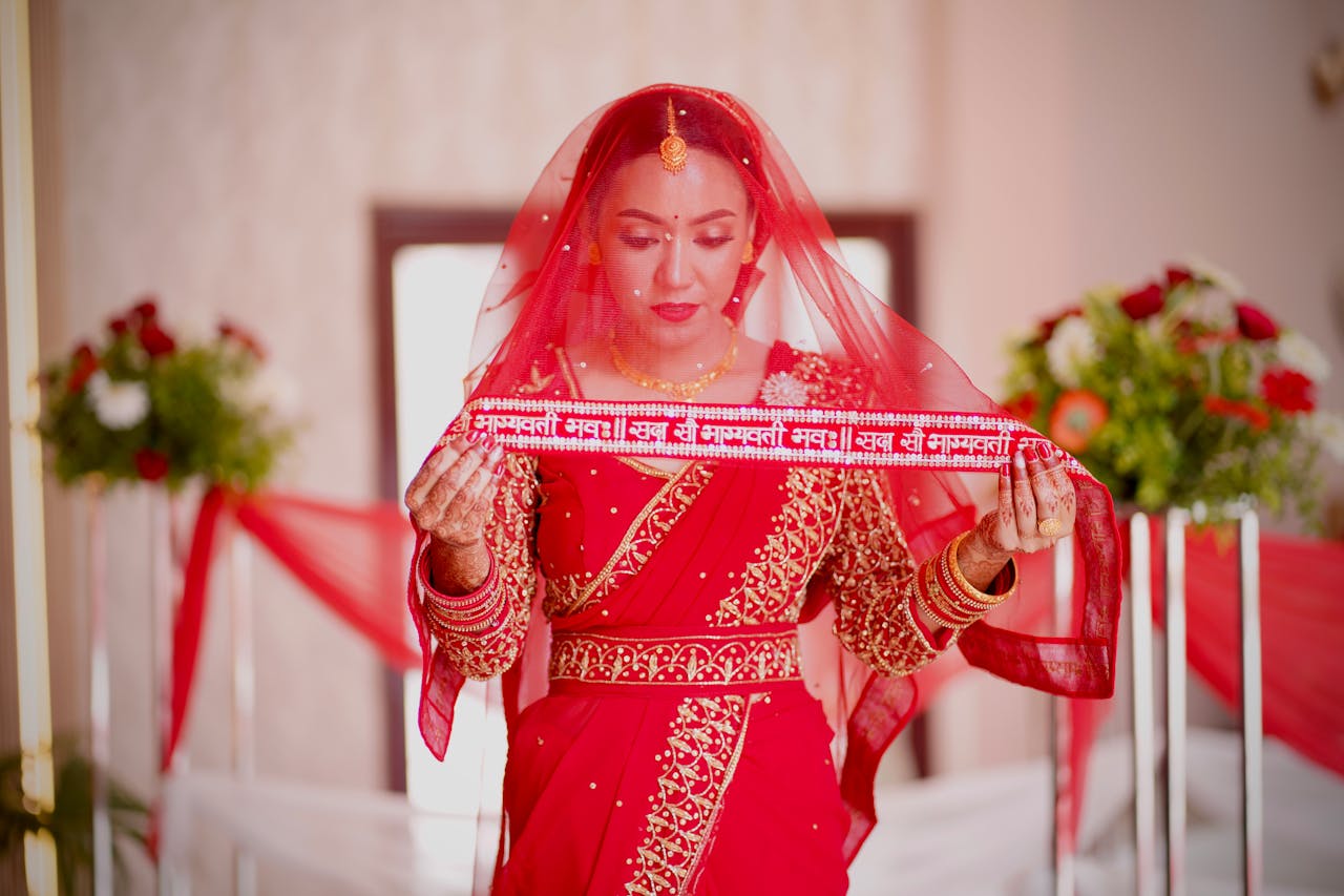 A beautiful Indian bride in red traditional attire, standing indoors with floral decor.