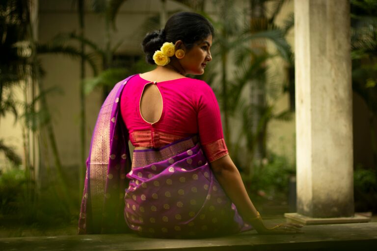 A back view of an Indian woman in traditional attire sitting outdoors, adorned with yellow flowers.